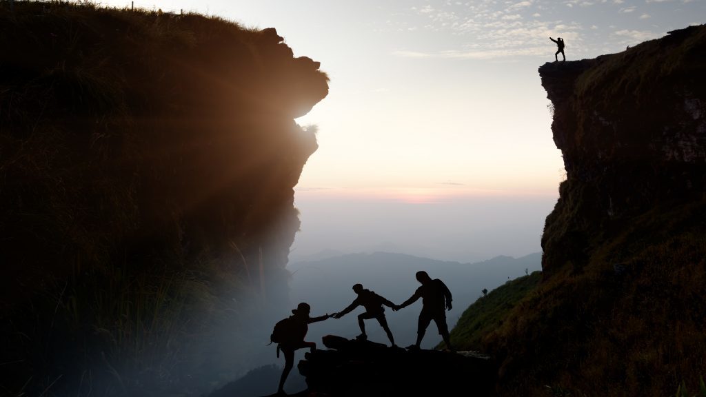 Mannwanderer, der den Erfolg auf einem Berg in einem majestätischen Sonnenaufgang feiert und Freunde der Klettergruppe helfen zu wandern.